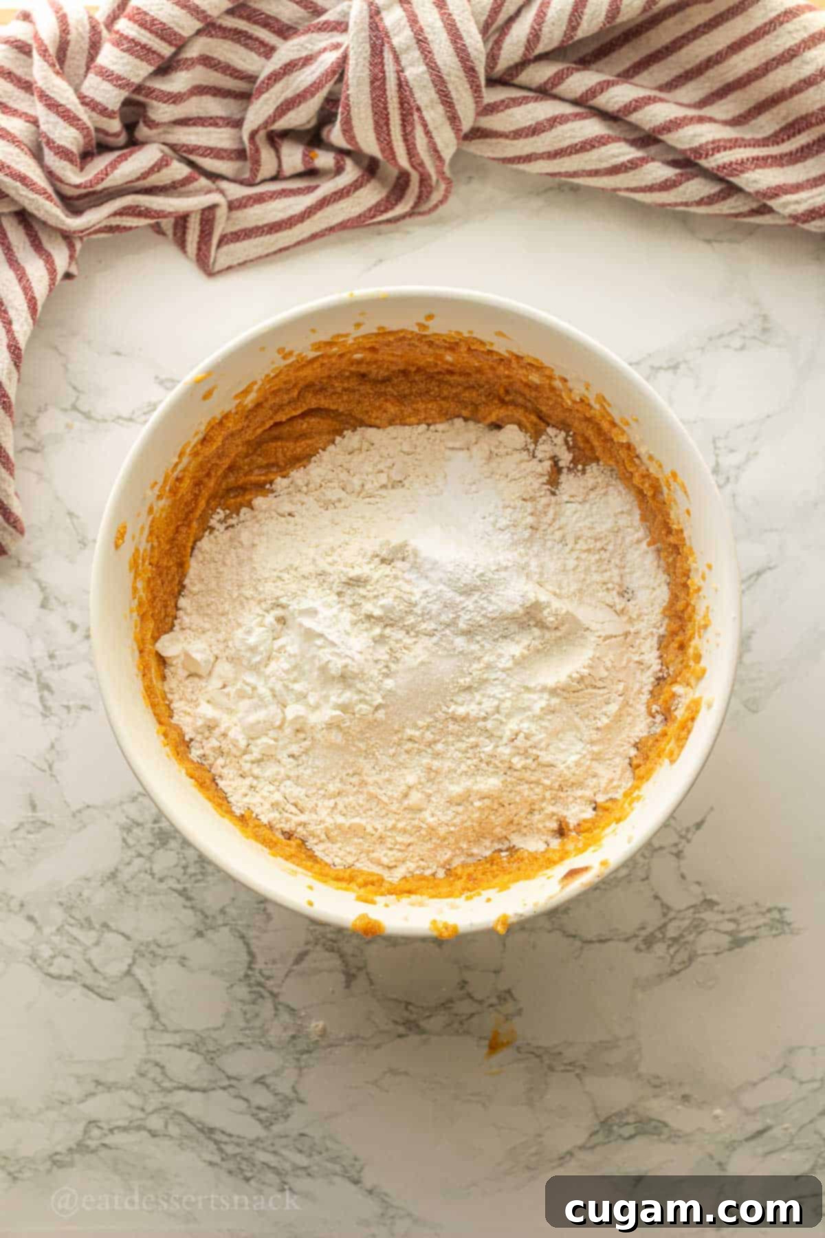 Flour, salt, baking soda, and baking powder on top of pumpkin cookie dough wet ingredients in bowl, ready for mixing.