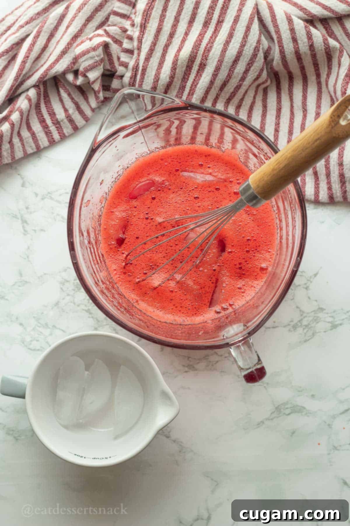 A whisk stirring ice cubes into the dissolved jello mixture in a glass bowl, rapidly cooling the gelatin base.